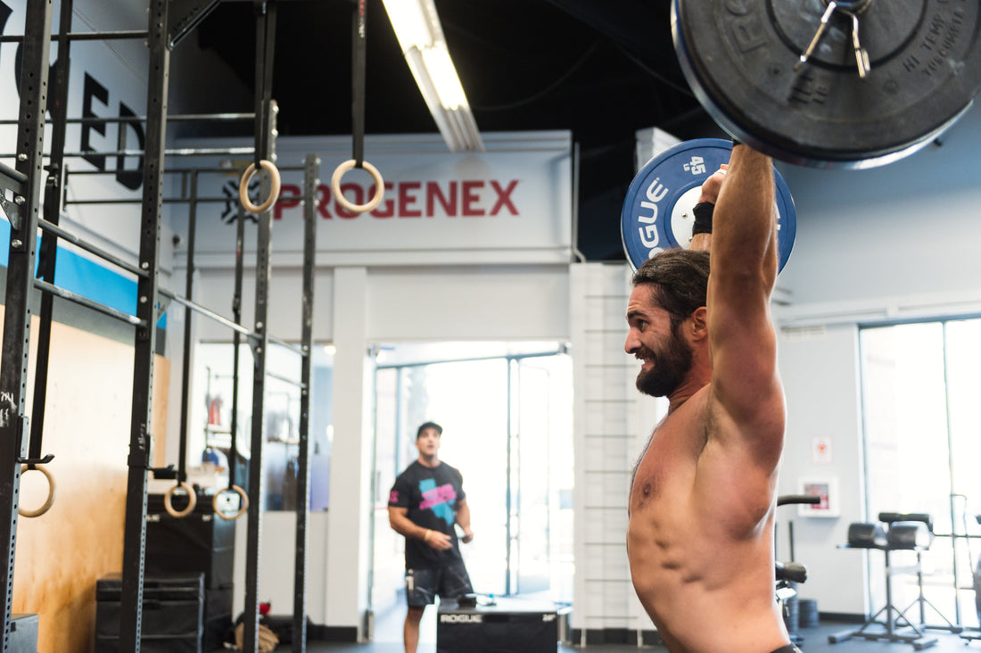 Shirtless man pressing a barbell overhead in a gym with a Progenex sign behind