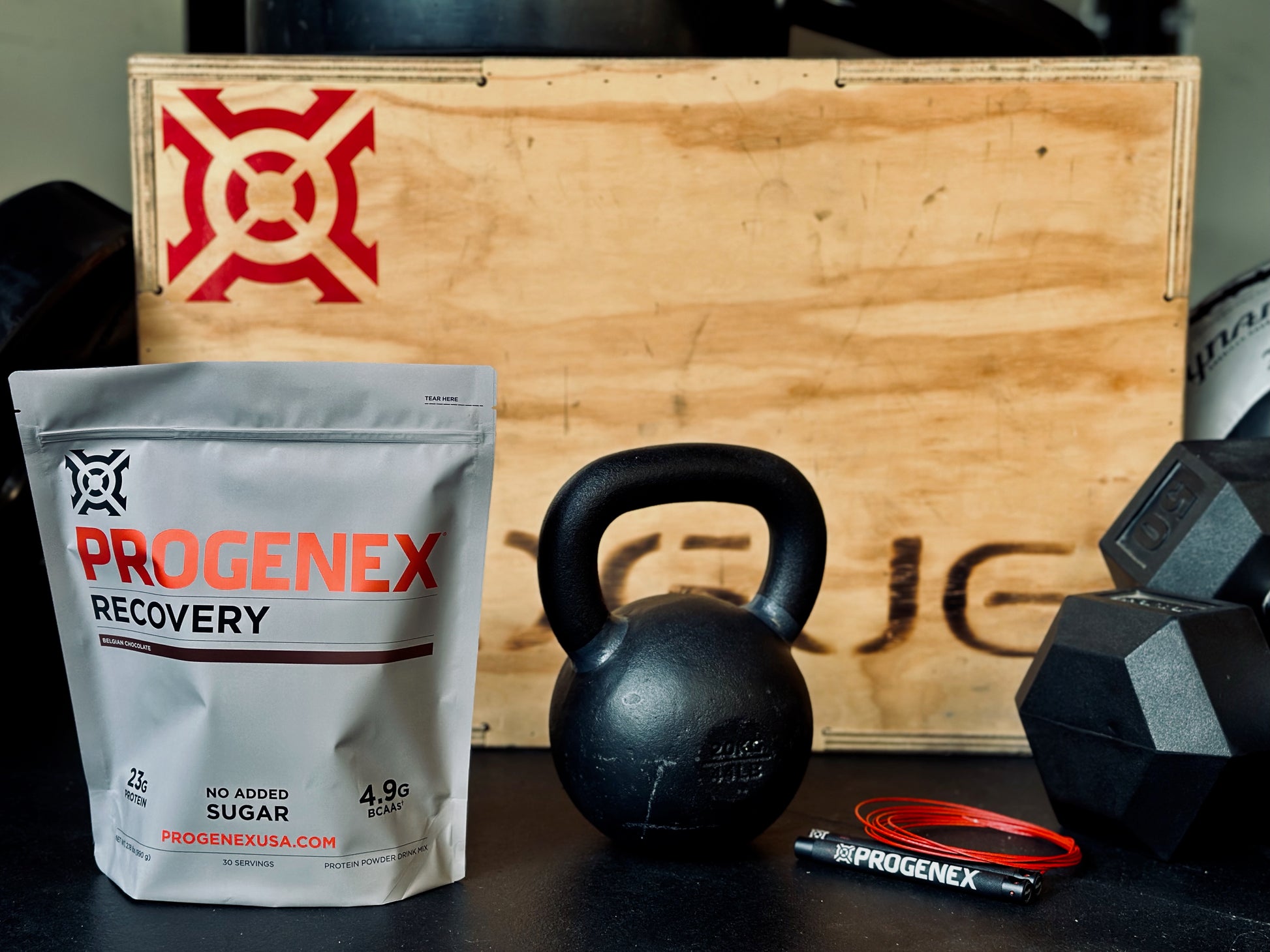 Progenex recovery supplement package with kettlebell and dumbbells on a wooden box