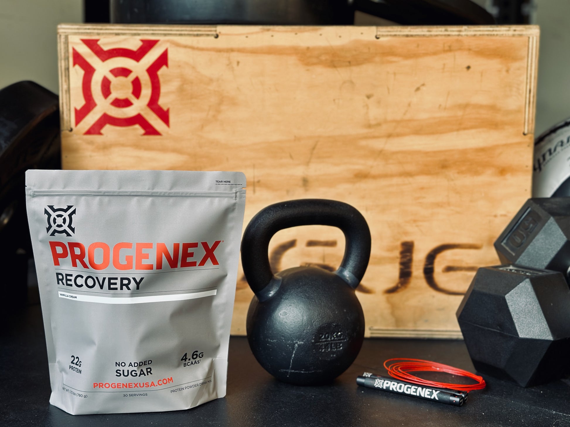 Progenex Recovery supplement package with kettlebell and dumbbells on a wooden box background